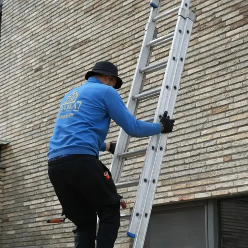 Dakdekker in blauwe trui klimt een ladder op tegen een bakstenen gevel in Beekbergen